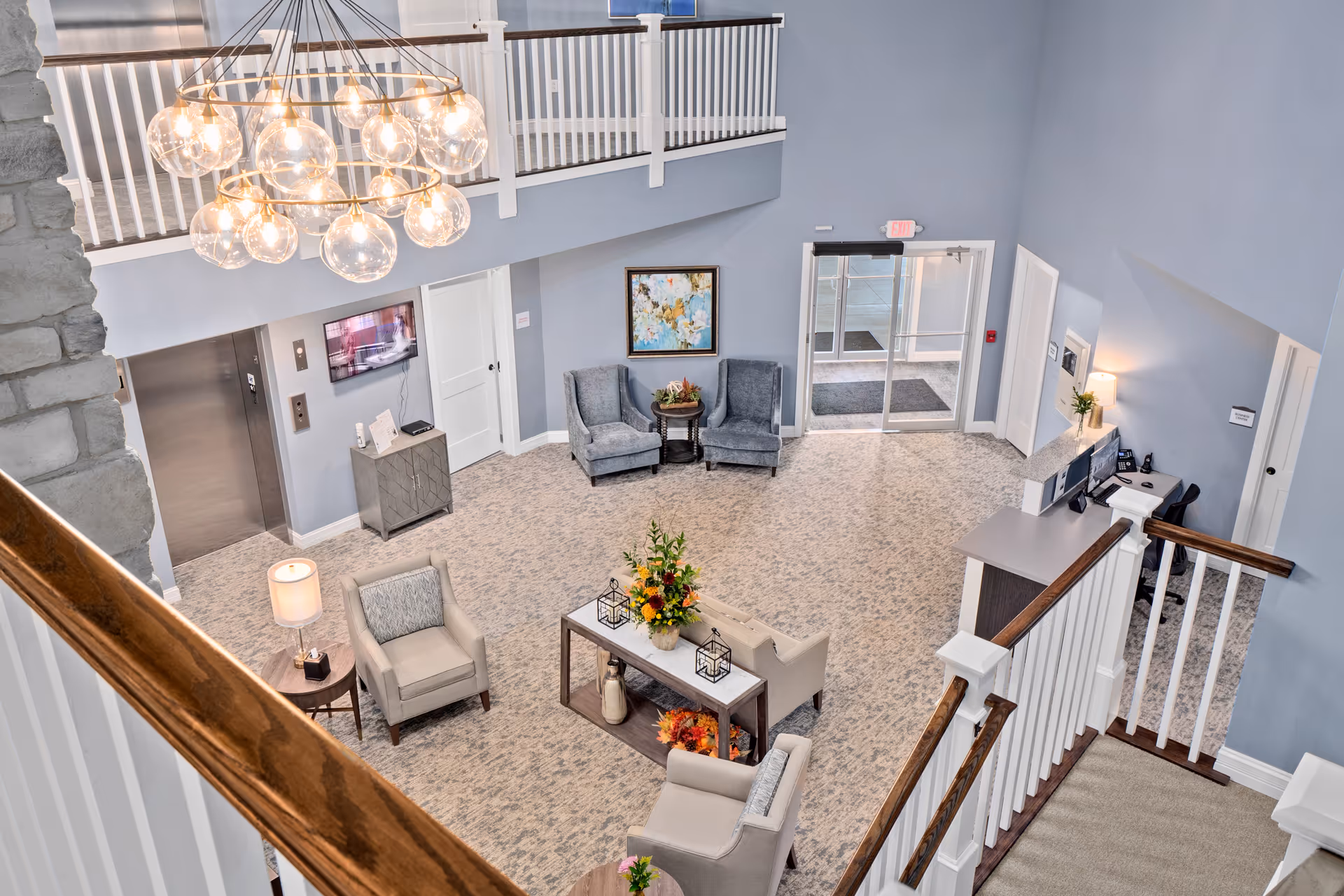 Bright, two-story senior living lobby with seating areas, a chandelier, reception desk, elevator and entrance doors.