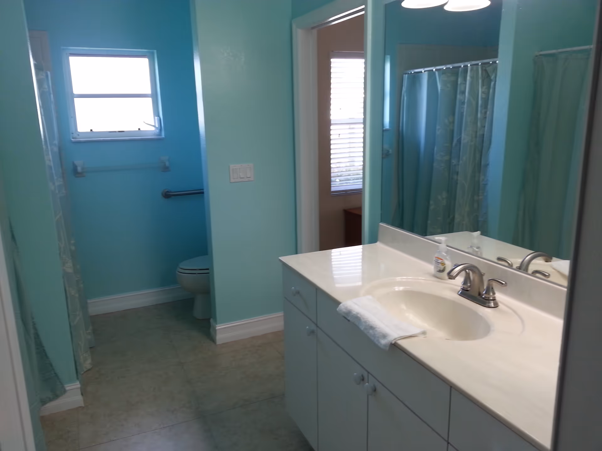 A bathroom with light blue walls featuring a white sink and countertop with a faucet and a hand towel. There is a mirror above the sink reflecting a shower with a blue floral shower curtain. A toilet is visible in a separate alcove with a small window above it.