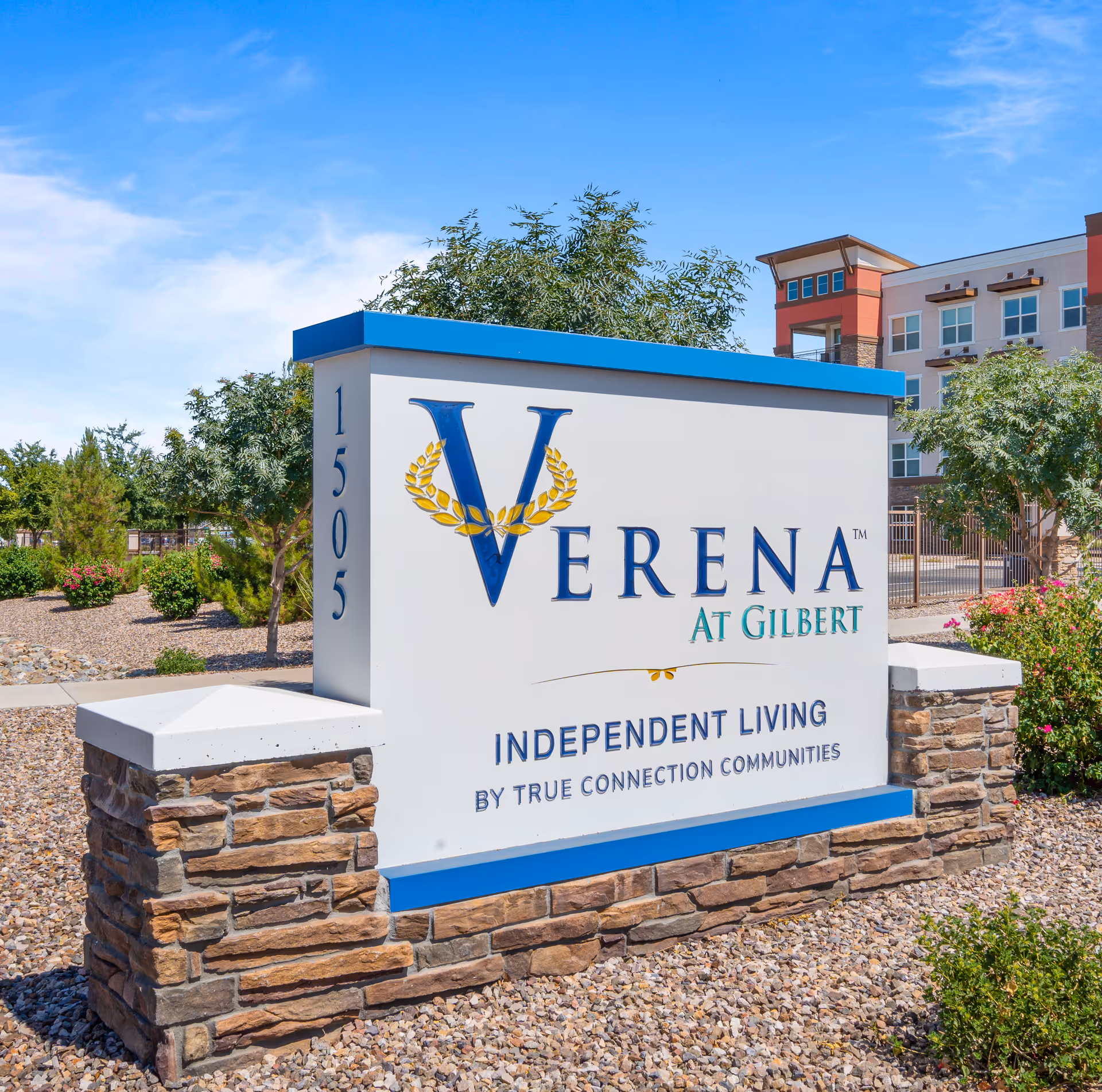 Outdoor view of a large sign for Verena at Gilbert, an independent living community by True Connection Communities, with a modern building and landscaping in the background under a clear blue sky.