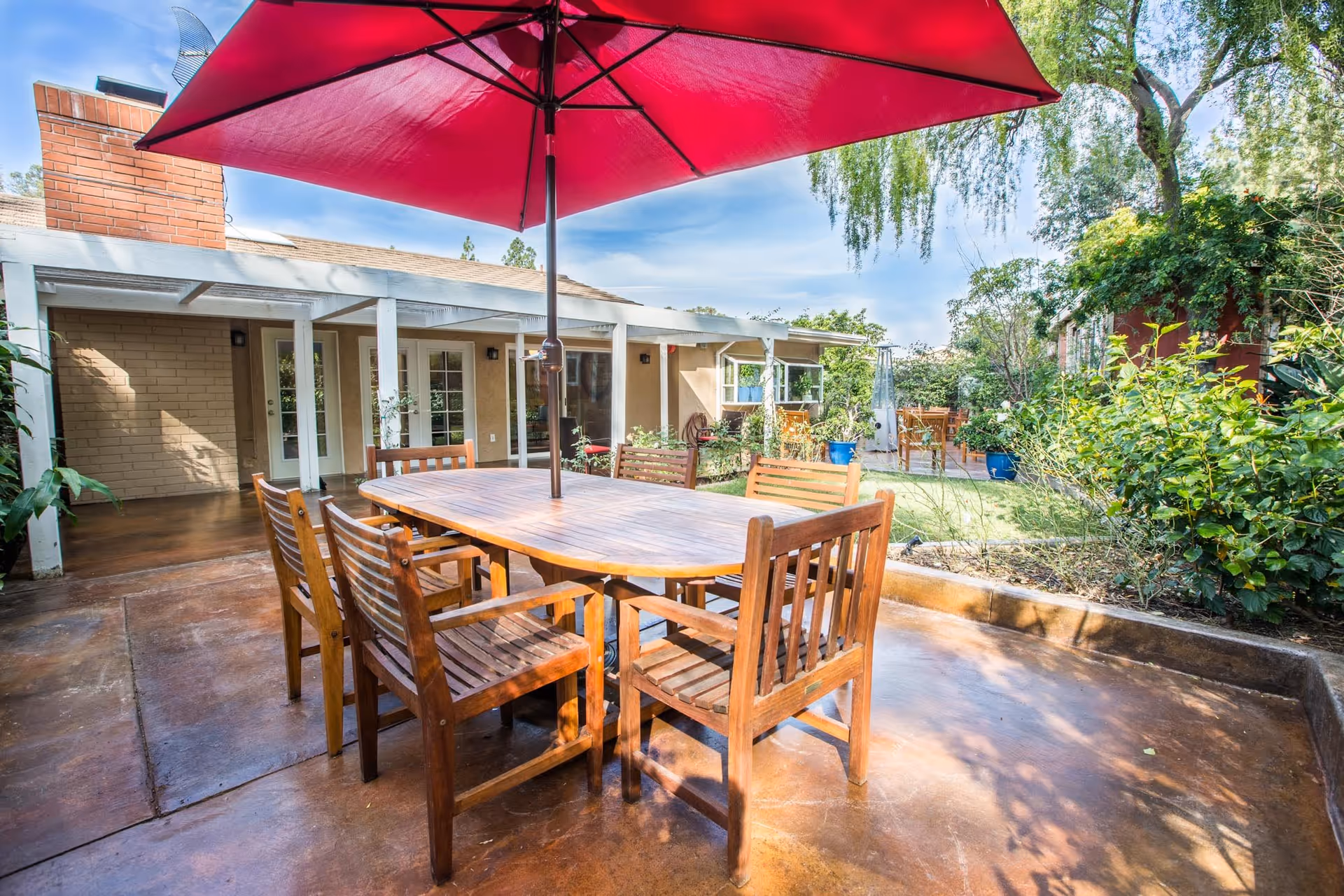 Outdoor patio area with a wooden table and six matching wooden chairs under a large red umbrella. The patio is adjacent to a building with beige walls and white-framed glass doors and windows. Surrounding the patio are green plants and trees under a partly cloudy blue sky.