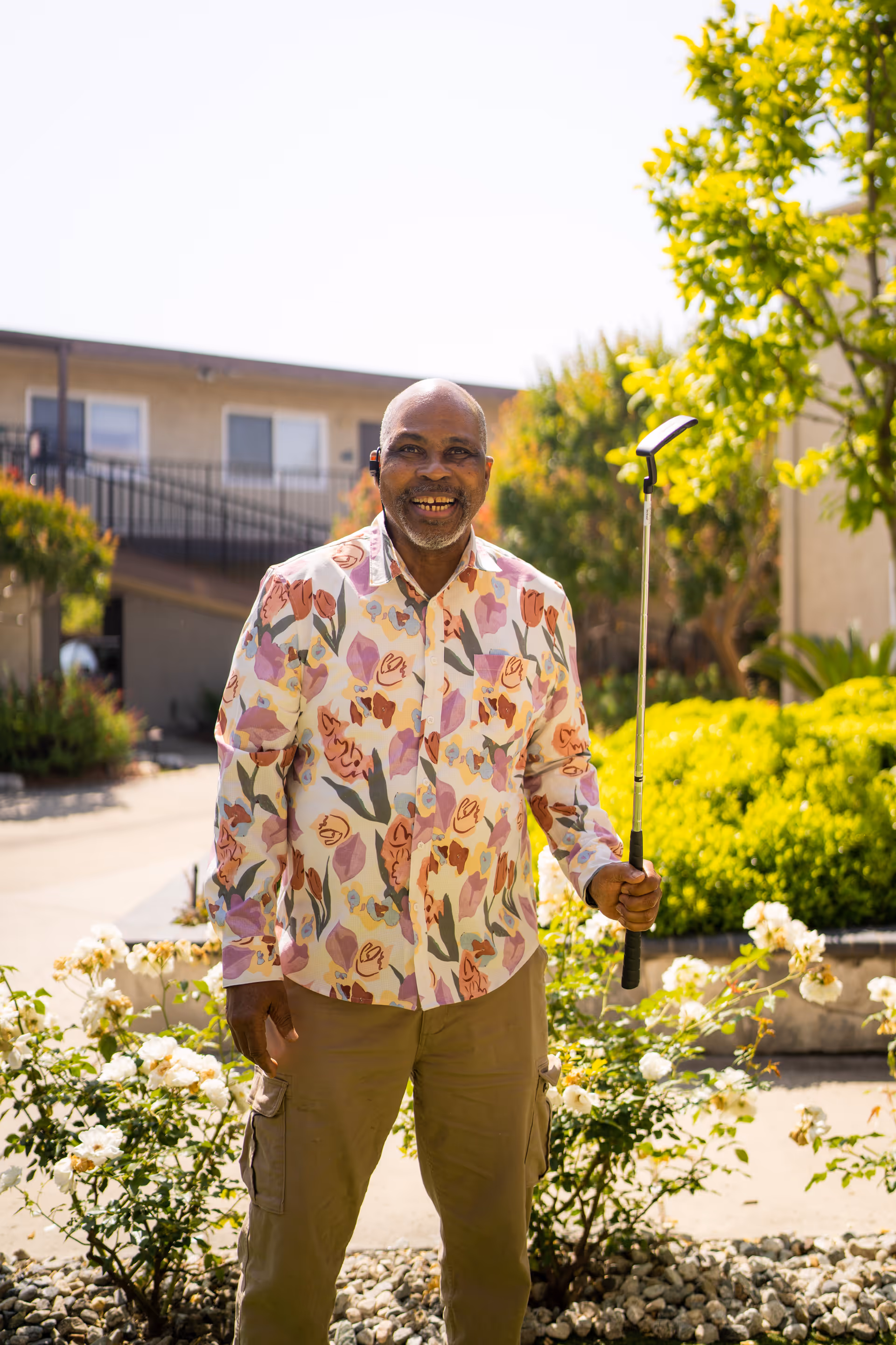 A smiling man standing outdoors in a garden area of a senior living facility, holding a golf club. He is wearing a colorful floral shirt and beige pants. Behind him are bushes, flowers, and a two-story building with windows and a staircase.