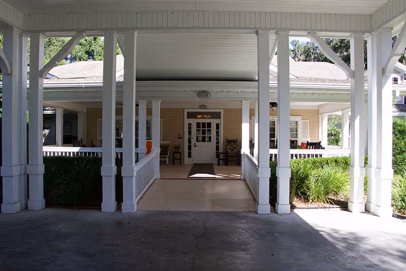 Covered porte-cochère with white columns leading to the porch and main entrance of a light-yellow building with seating.