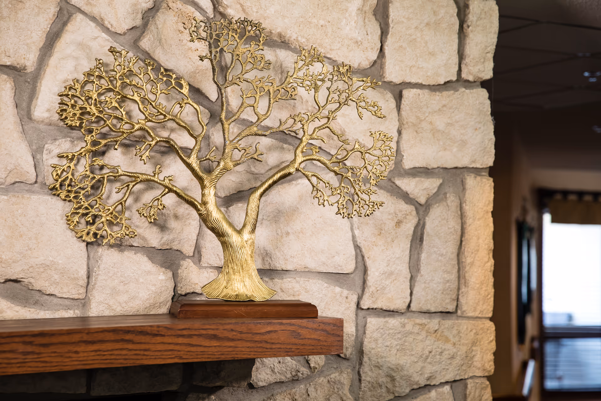 Decorative brass tree sculpture on a wooden mantel against a stone fireplace wall in a common living area.