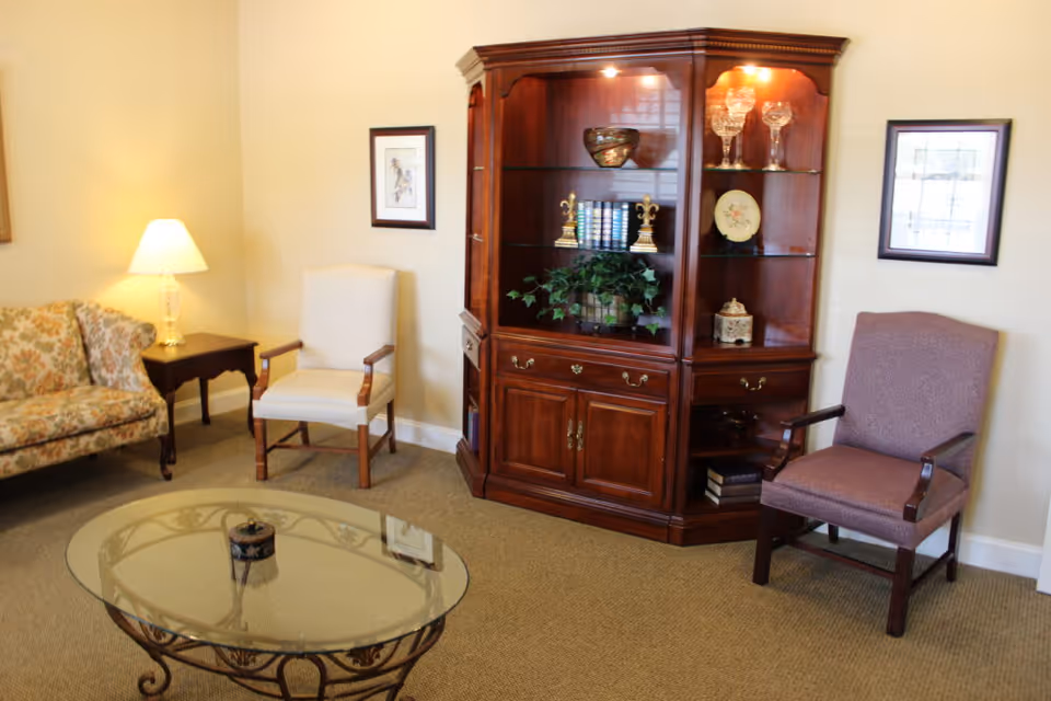 A cozy living room area with a floral patterned sofa, two armchairs, a wooden side table with a lamp, a glass coffee table with a decorative item, and a wooden display cabinet holding books, glassware, and decorative pieces. The walls are light-colored with framed artwork.