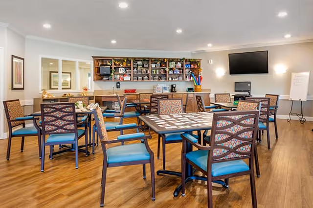 A bright and spacious common area with multiple tables and chairs arranged for group activities. The tables have card games set up on them. In the background, there is a counter with shelves holding various items, a microwave, and a coffee maker. A large flat-screen TV is mounted on the wall, and a whiteboard on an easel stands nearby. The room has wood flooring and recessed lighting in the ceiling.