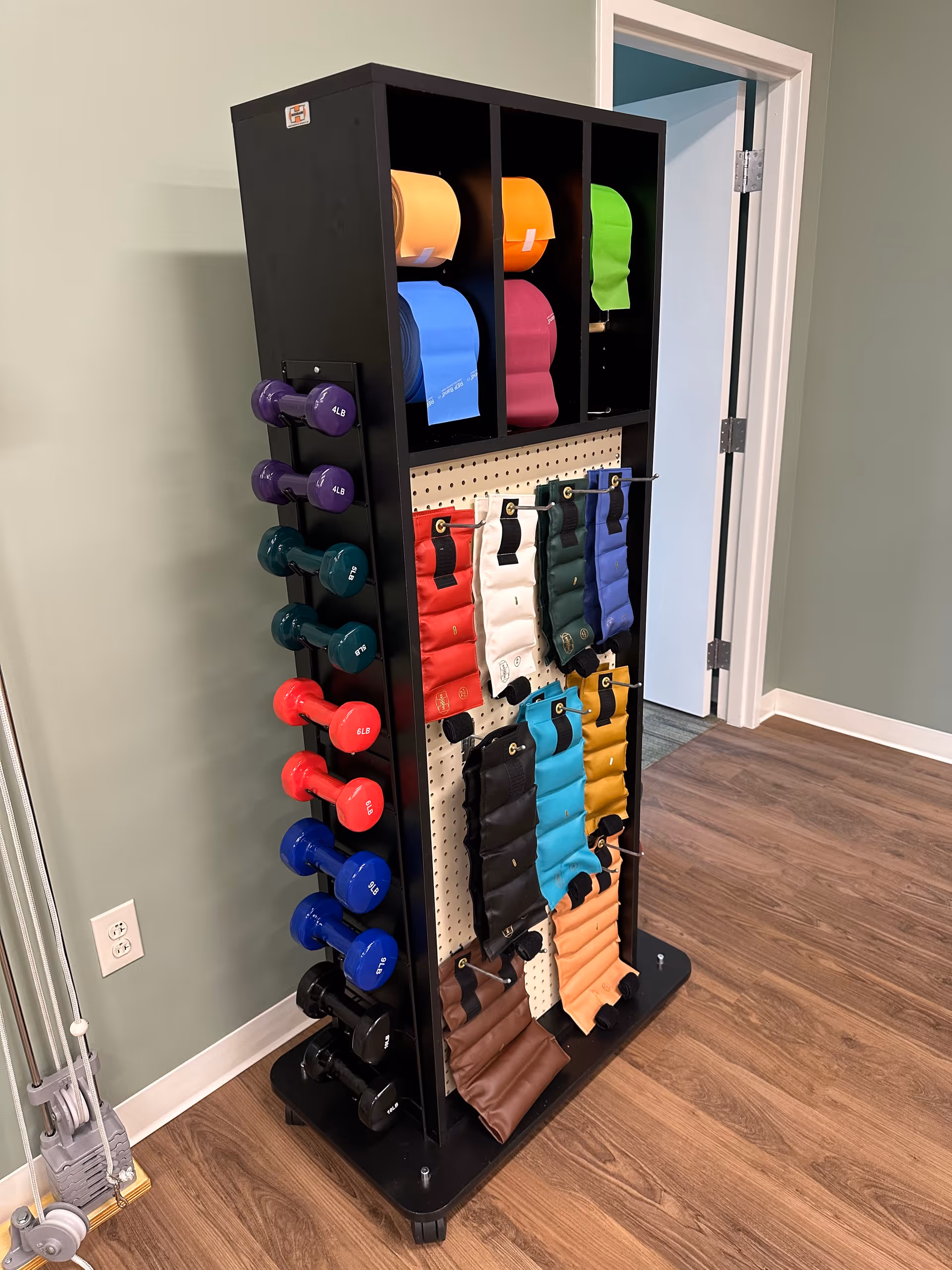 A tall equipment rack holding colorful dumbbells, resistance bands, and weighted straps in a room with wood flooring and an open doorway.