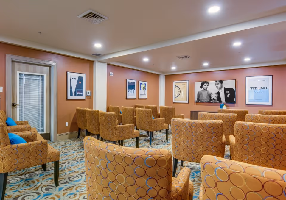 A cozy meeting or lounge room with multiple patterned orange armchairs arranged in rows facing a wall with framed black and white and color artwork. The room has warm orange walls, a patterned carpet, and recessed ceiling lights. There is a door with a window on the left side of the room.