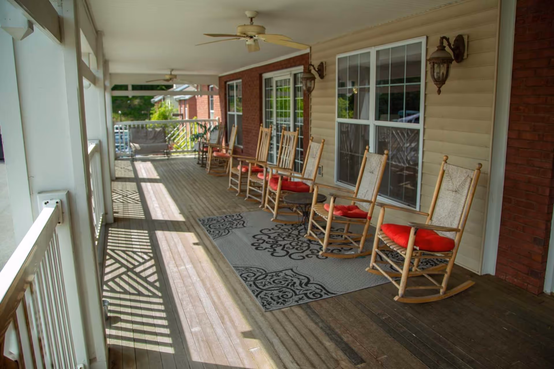 A covered outdoor porch area with several wooden rocking chairs with red cushions arranged along the wall. The porch has ceiling fans, wall-mounted lantern-style lights, a patterned rug, and a wooden floor. The porch overlooks a green area and some buildings in the background.