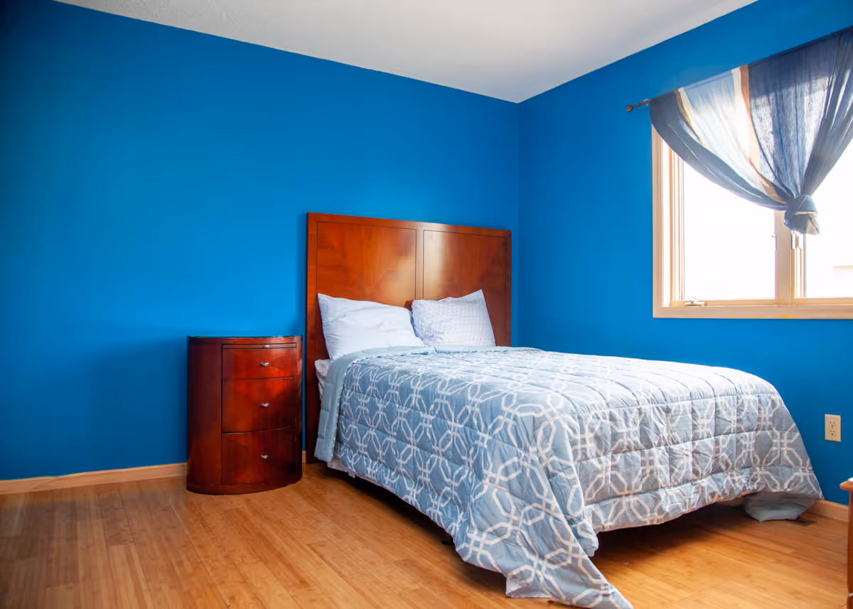 A bedroom with bright blue walls, a wooden bed frame with a patterned blue and white comforter, two pillows, a wooden nightstand with three drawers, and a window with a sheer blue curtain tied in the middle letting in natural light.