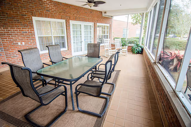 A covered outdoor patio area with a glass-top table surrounded by six black mesh chairs on a brown rug. The patio has a brick wall with windows and a door, two white rocking chairs, a ceiling fan, and large screened windows overlooking greenery outside.