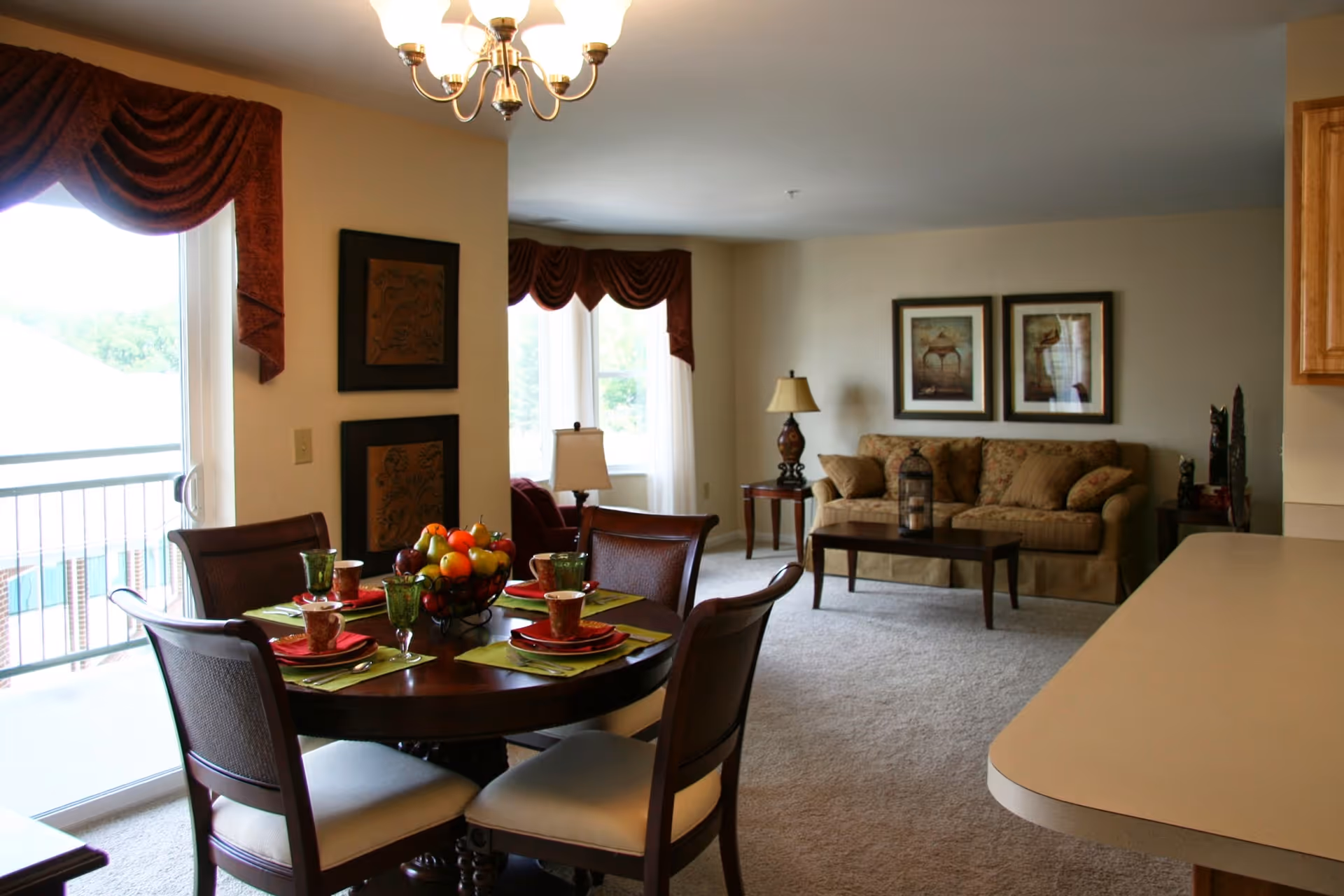 Interior view of a senior living facility showing a dining area with a round table set for four with green placemats, red plates, cups, and a centerpiece of fruit. Adjacent to the dining area is a living room with a beige sofa, two framed pictures on the wall, a wooden coffee table, and a side table with a lamp. Large windows with red valances allow natural light into the space.