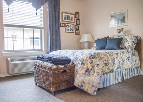 A cozy bedroom in Oak Tree Assisted Living featuring a bed with floral bedding and blue pillows, a wicker storage chest at the foot of the bed with folded blankets on top, a window with blinds and blue curtains, a white table lamp on a nightstand, and framed pictures on the wall.