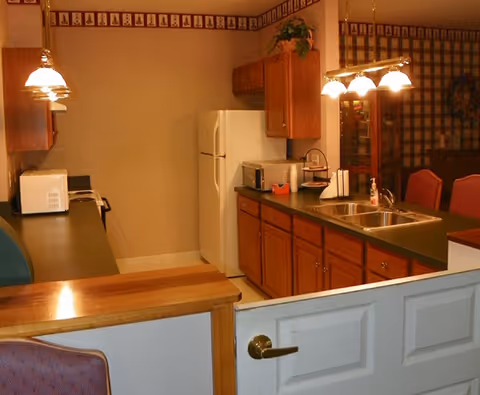 Interior view of a kitchen area in a senior living facility featuring wooden cabinets, a white refrigerator, a microwave, a double sink, and hanging light fixtures. The kitchen counter extends into a dining area with red cushioned chairs and a glass-front cabinet in the background.