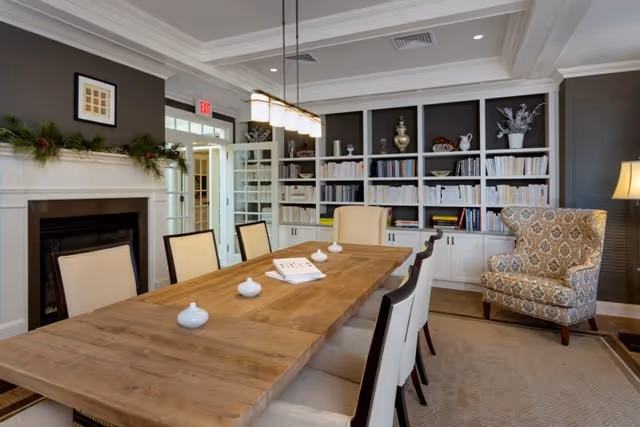 A well-lit interior room featuring a long wooden table surrounded by cushioned chairs. The room has a built-in bookshelf filled with books and decorative items, a patterned armchair, a fireplace with a garland on the mantle, and a hanging light fixture above the table. The walls are painted gray with white trim and coffered ceiling details.