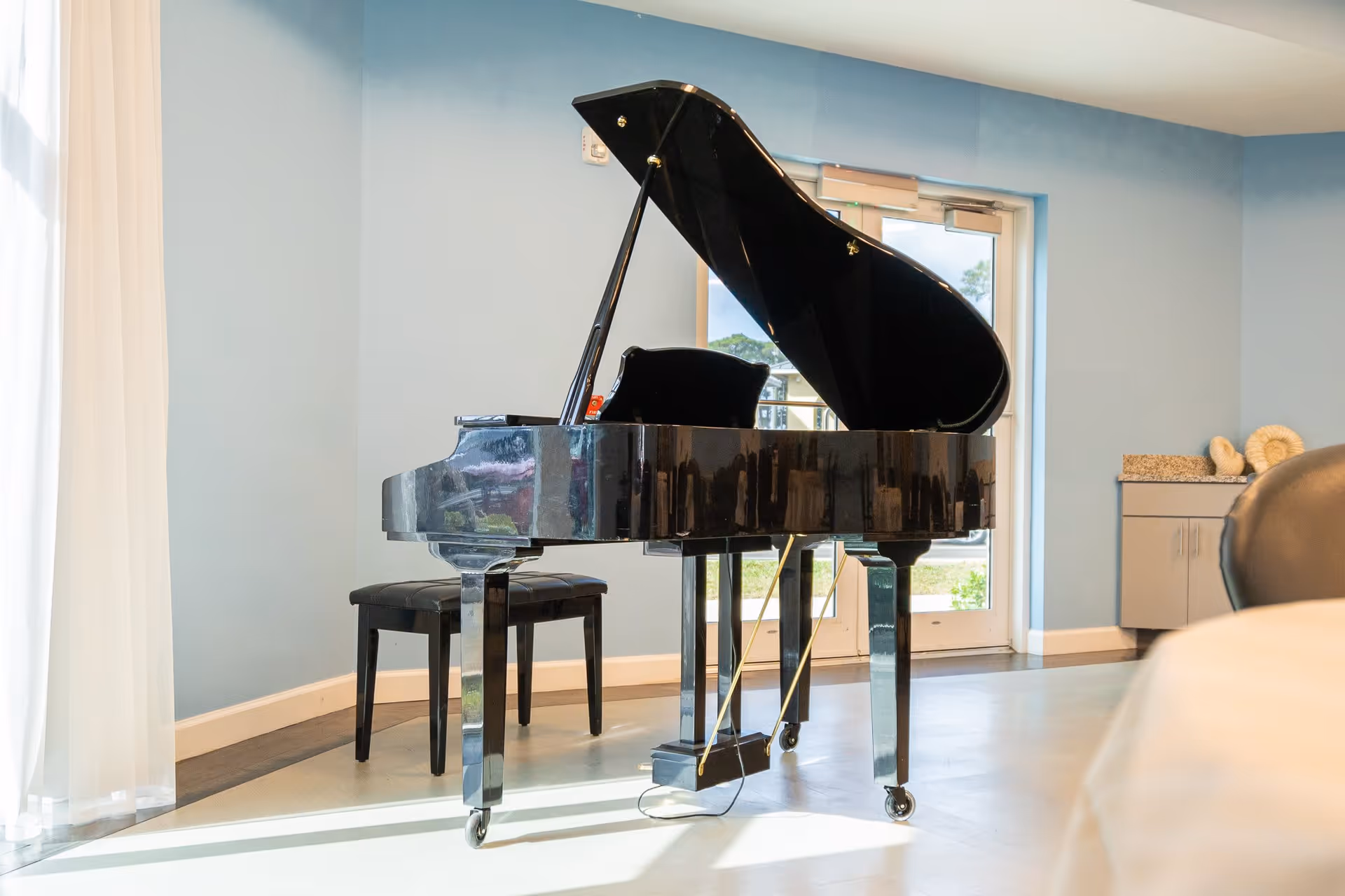 A black grand piano with its lid open is positioned in a bright room with light blue walls and a large window with sheer white curtains. There is a black piano bench in front of the piano and a cabinet with decorative items on the right side near a glass door leading outside.