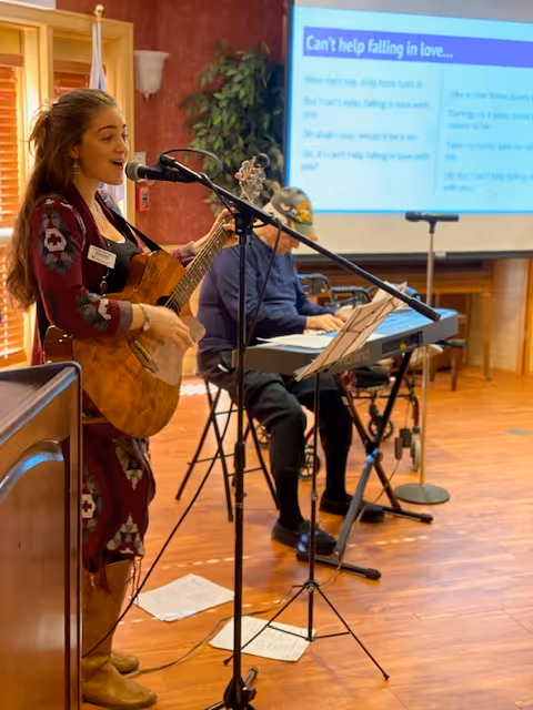 A musician sings and plays guitar while another plays a keyboard in a common room with song lyrics projected on a screen.
