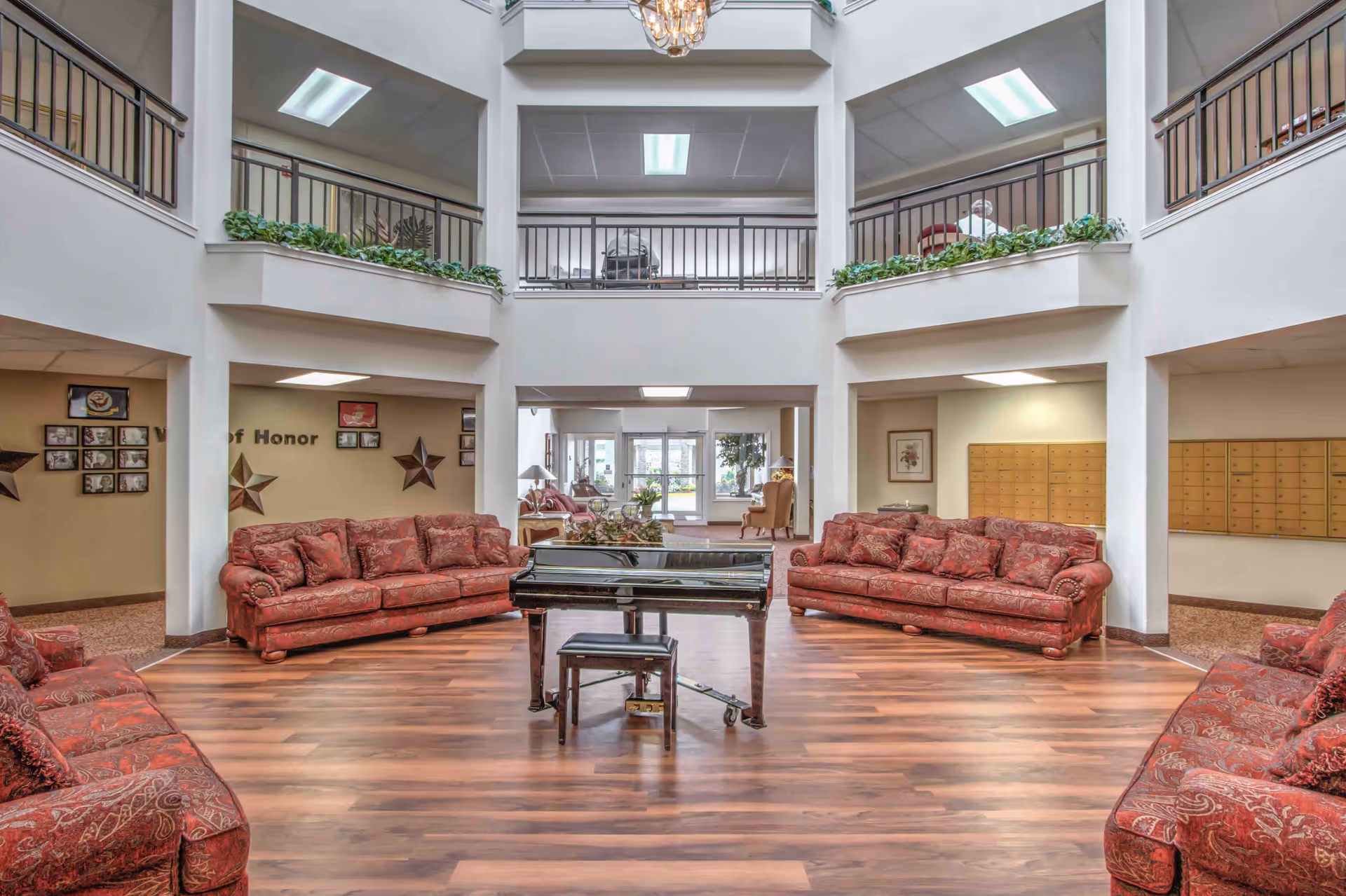 A spacious senior living facility common area with four red patterned sofas arranged around a black grand piano on a wooden floor. The room has high ceilings with an upper balcony railing and a chandelier hanging from above. There are plants on the balcony ledges and framed pictures on the walls.