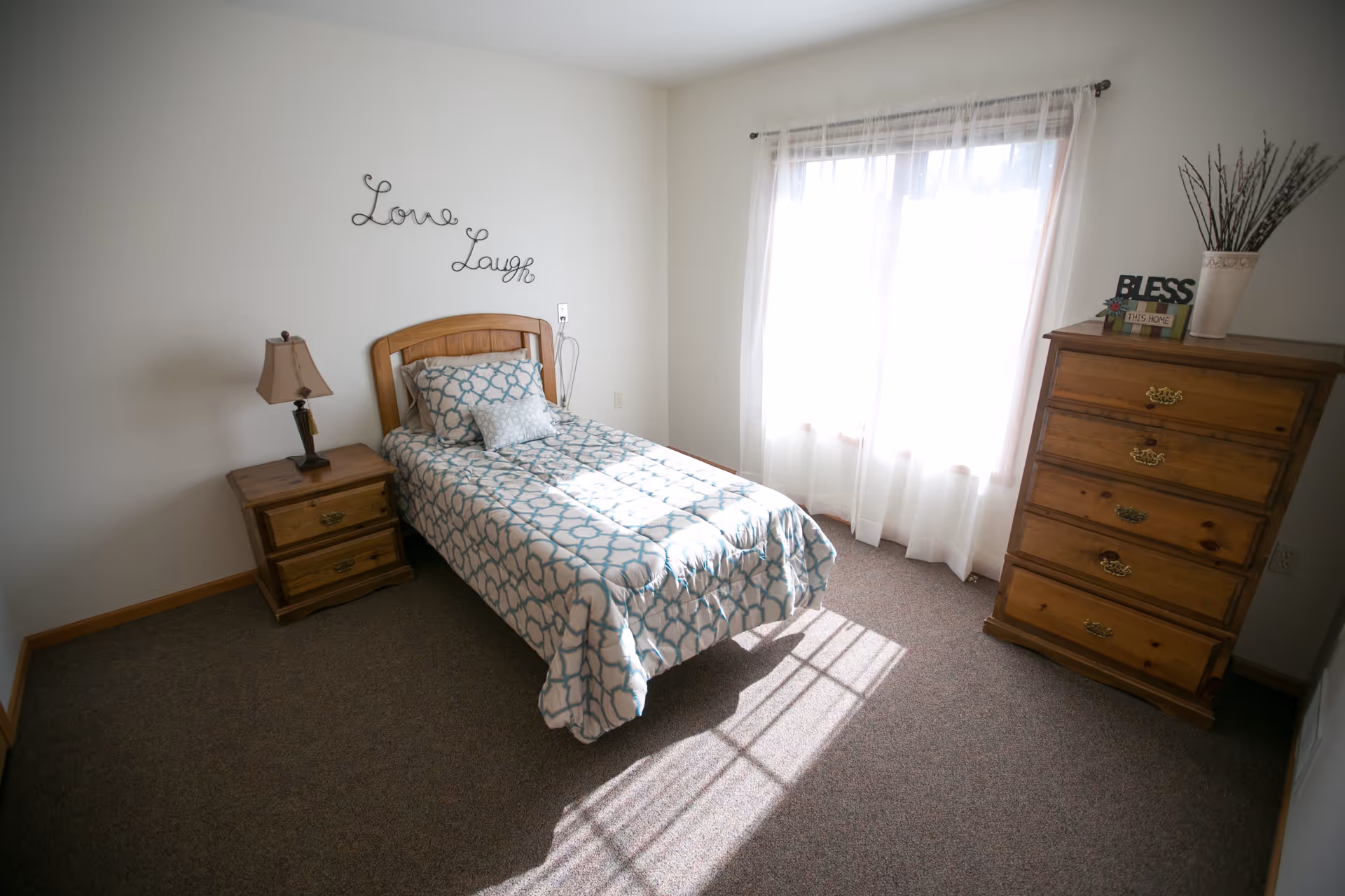 A bright bedroom with a single bed covered in a patterned blue and white comforter, two pillows, and a wooden headboard. There is a wooden nightstand with a lamp on the left side of the bed. On the right side of the room, there is a wooden dresser with decorative items including a vase with branches and a sign that says 'BLESS THIS HOME'. Sheer white curtains cover a large window letting in sunlight. The wall above the bed has decorative metal words that say 'Love Laugh'.