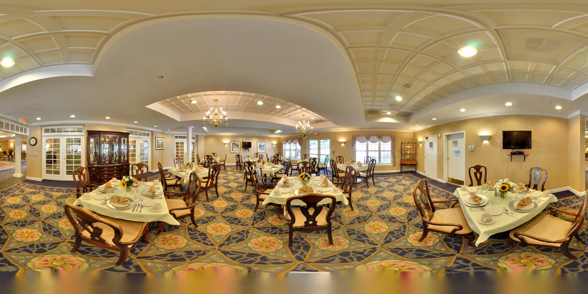 A spacious senior living dining room with multiple tables set for meals. Each table has a white tablecloth, neatly folded napkins, plates, cups, and silverware. The room features patterned carpet, chandeliers, large windows with curtains, and a wall-mounted television. There is a china cabinet and decorative wall art, creating a warm and inviting atmosphere.