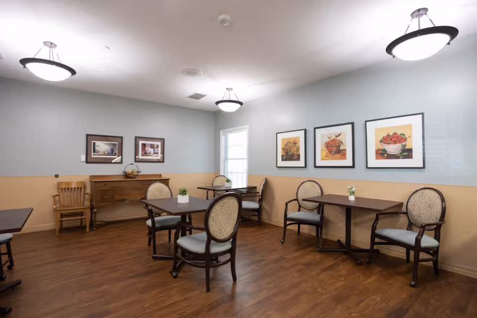 Small communal dining room with tables, upholstered chairs, framed artwork, and pendant lights.
