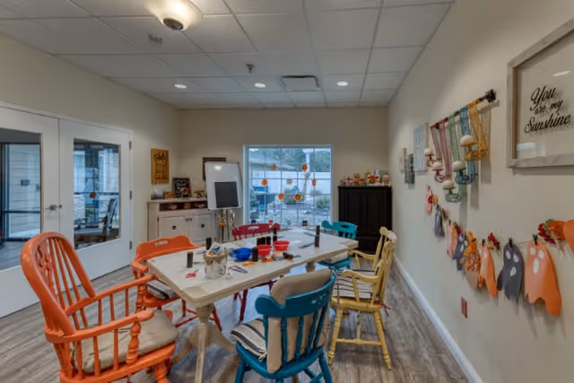 A bright and colorful activity room in a senior living facility with a rectangular table surrounded by mismatched chairs in orange, blue, yellow, and red. The table has various craft supplies on it. The walls are decorated with cheerful artwork and hanging crafts. A window lets in natural light, and double glass doors lead to another room.