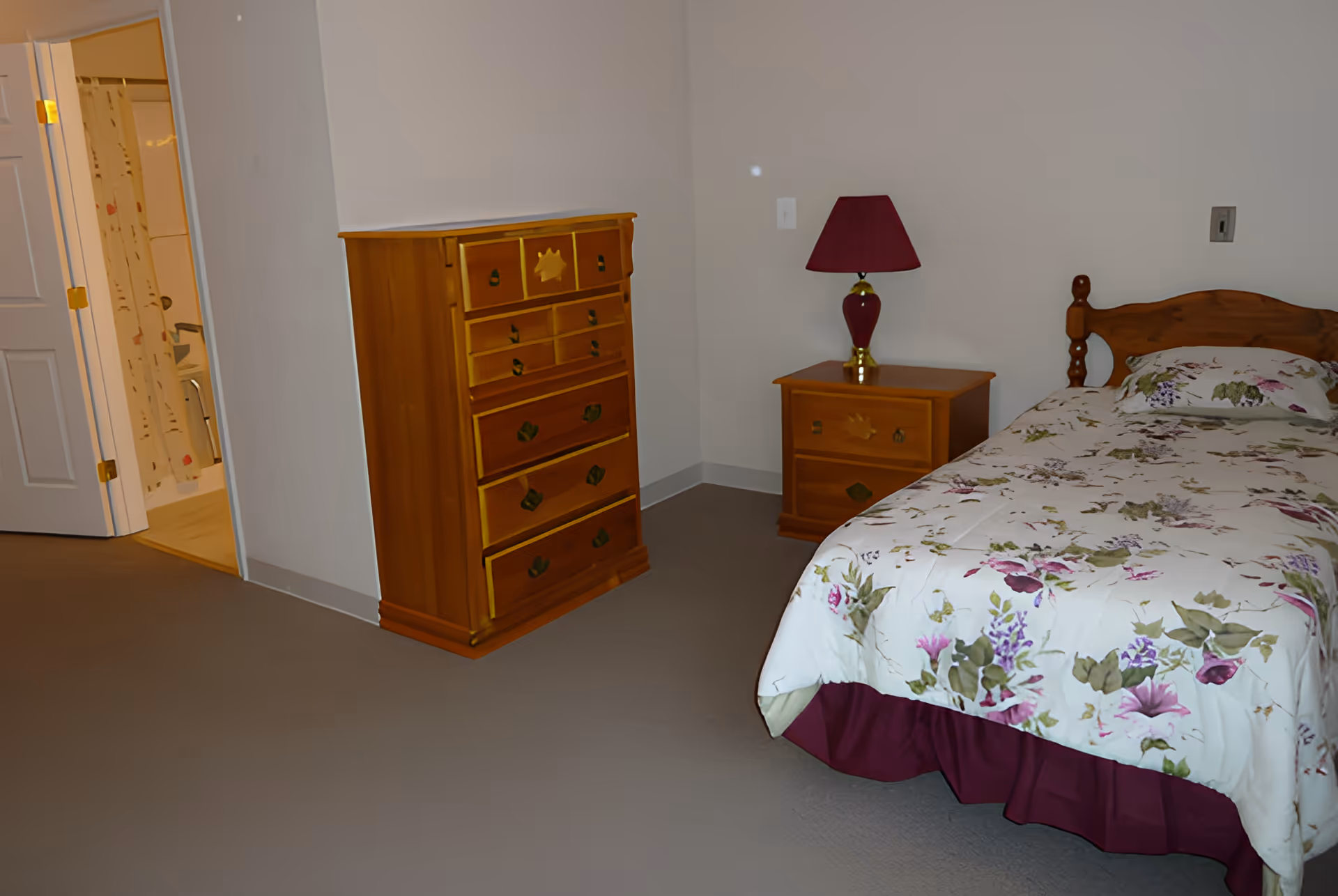 Small bedroom with a single bed dressed in floral bedding, a wooden dresser and nightstand with a lamp, and an open door to a bathroom.