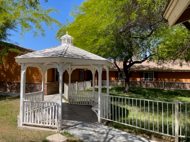 White wooden gazebo on a paved walkway in a shaded courtyard with grass and a single-story building in the background.