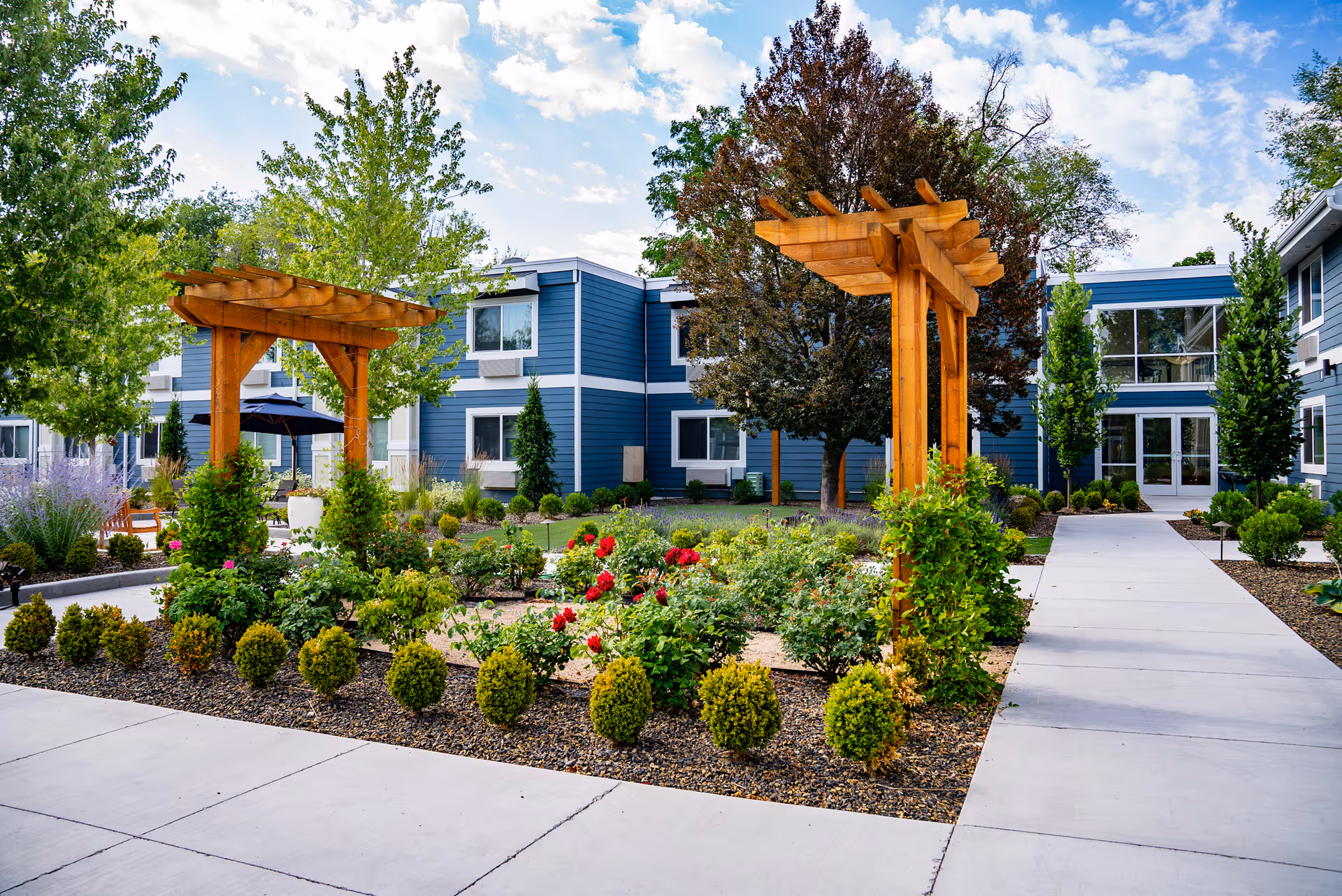 Outdoor garden area at Heatherwood Senior Living featuring a landscaped flower bed with red and pink flowers, small bushes, and two wooden pergolas. The garden is surrounded by paved walkways and trees, with a blue two-story building in the background under a partly cloudy sky.