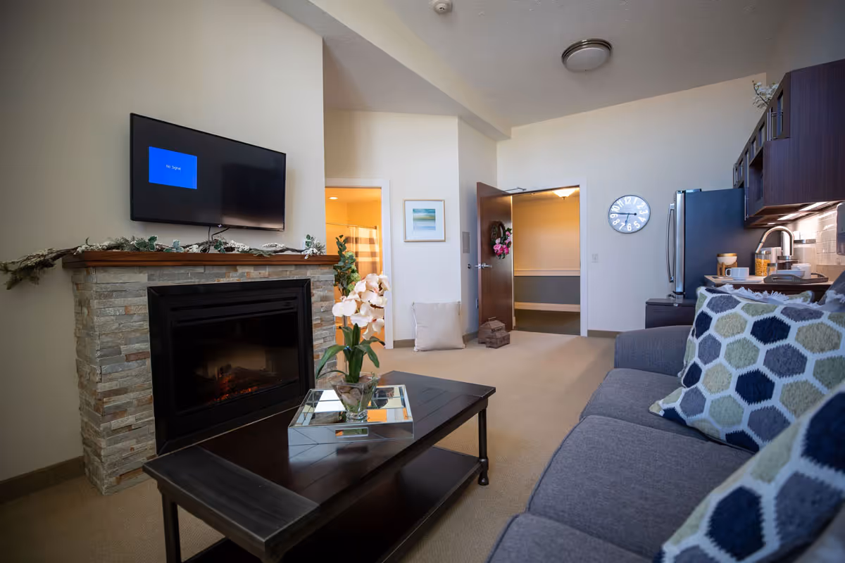 A cozy living room area in The Cortland Riverside Gardens featuring a stone fireplace with a mounted flat-screen TV above it showing a 'No Signal' message. In front of the fireplace is a dark wooden coffee table with a glass vase holding white orchids. To the right, there is a gray sofa with patterned cushions. The room has beige carpet and walls, with a kitchen area visible on the right side, including a refrigerator and cabinets. Two doorways are visible in the background, one leading to a bathroom with a striped shower curtain and the other to a hallway with a clock on the wall.