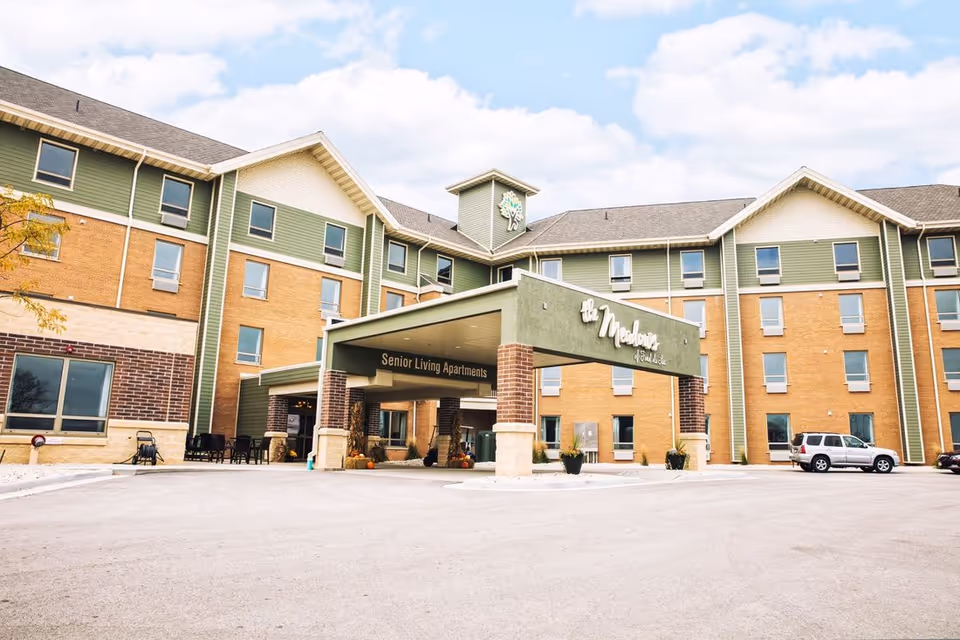 Exterior view of The Meadows Of Fond Du Lac senior living apartments building with a covered entrance and a few parked cars in front under a partly cloudy sky.