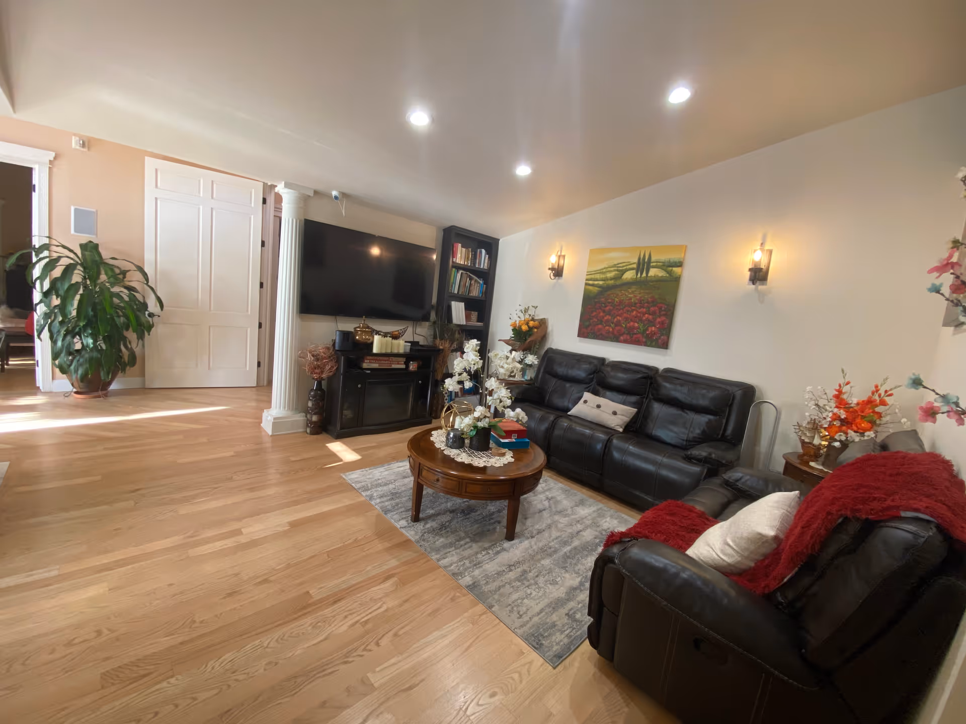 A cozy living room with light wood flooring, a black leather sofa and armchair with a red throw blanket, a round wooden coffee table with decorative items, a flat-screen TV mounted on the wall above a black fireplace, a bookshelf filled with books, wall sconces providing warm lighting, and a colorful landscape painting on the wall.