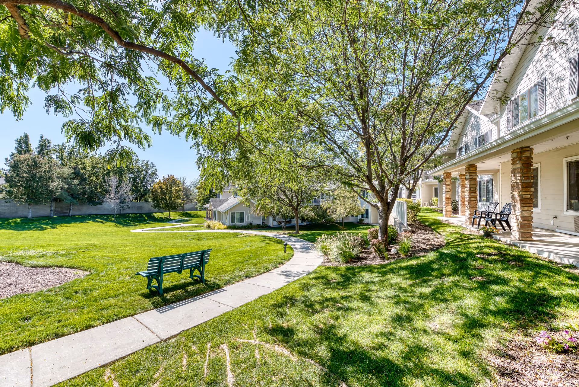 A sunny outdoor area at Solstice Senior Living at Sandy featuring a green lawn, a curved concrete walkway, a green bench, trees providing shade, and a building with a porch and stone pillars on the right side.