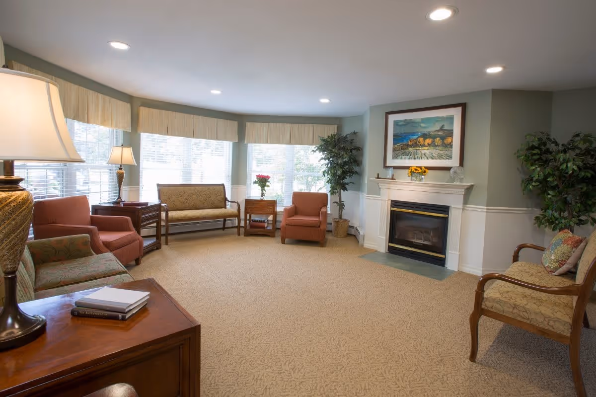 A cozy senior living room with beige carpet, several upholstered chairs and a loveseat arranged around a fireplace. The room has large windows with blinds and valances, two potted plants, a side table with a lamp, and a framed painting above the fireplace.