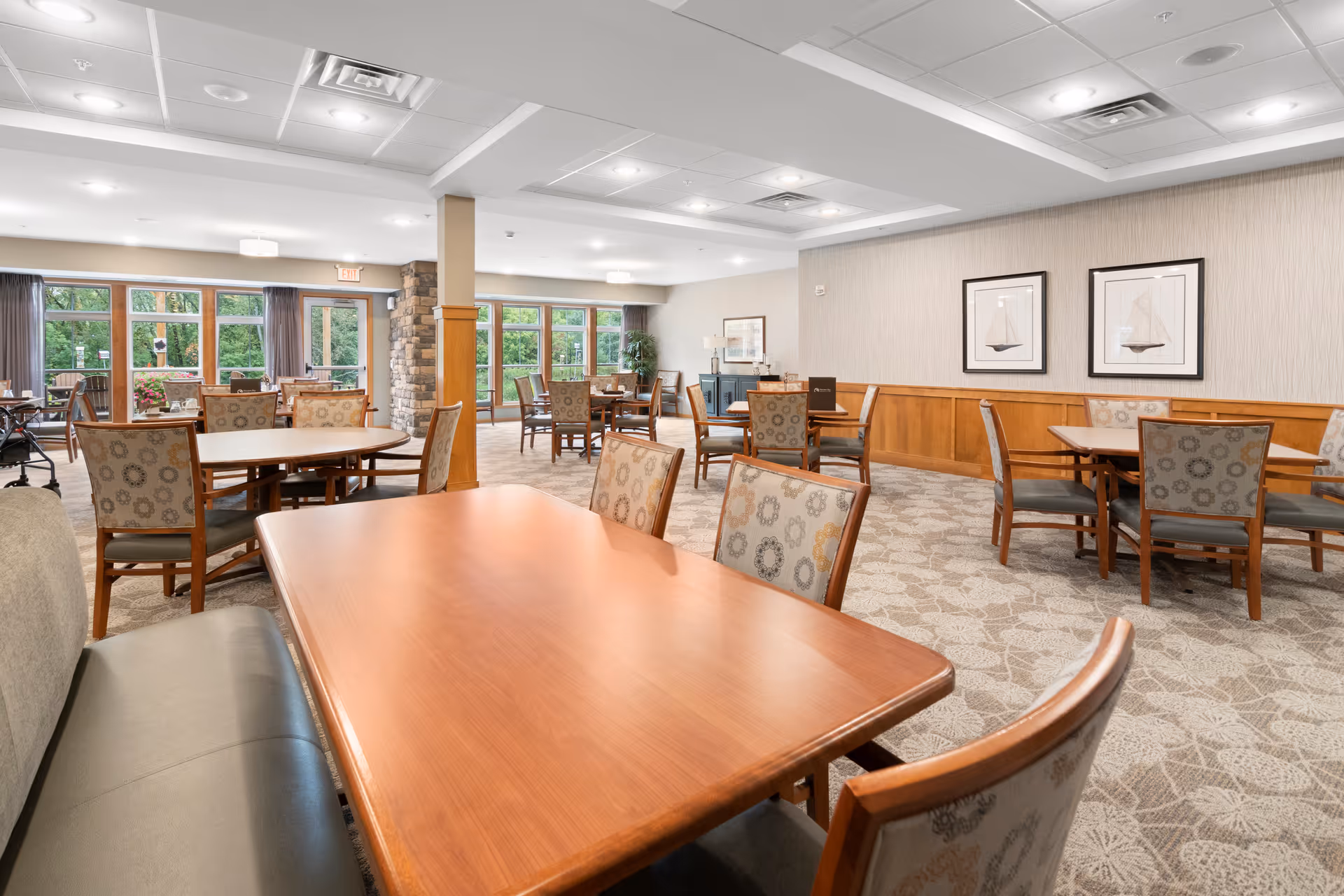A spacious senior living facility dining room with multiple wooden tables and cushioned chairs arranged neatly. Large windows along the walls allow natural light to brighten the room, and framed artwork featuring sailboats hangs on the wall. The room has a carpeted floor and a ceiling with recessed lighting.