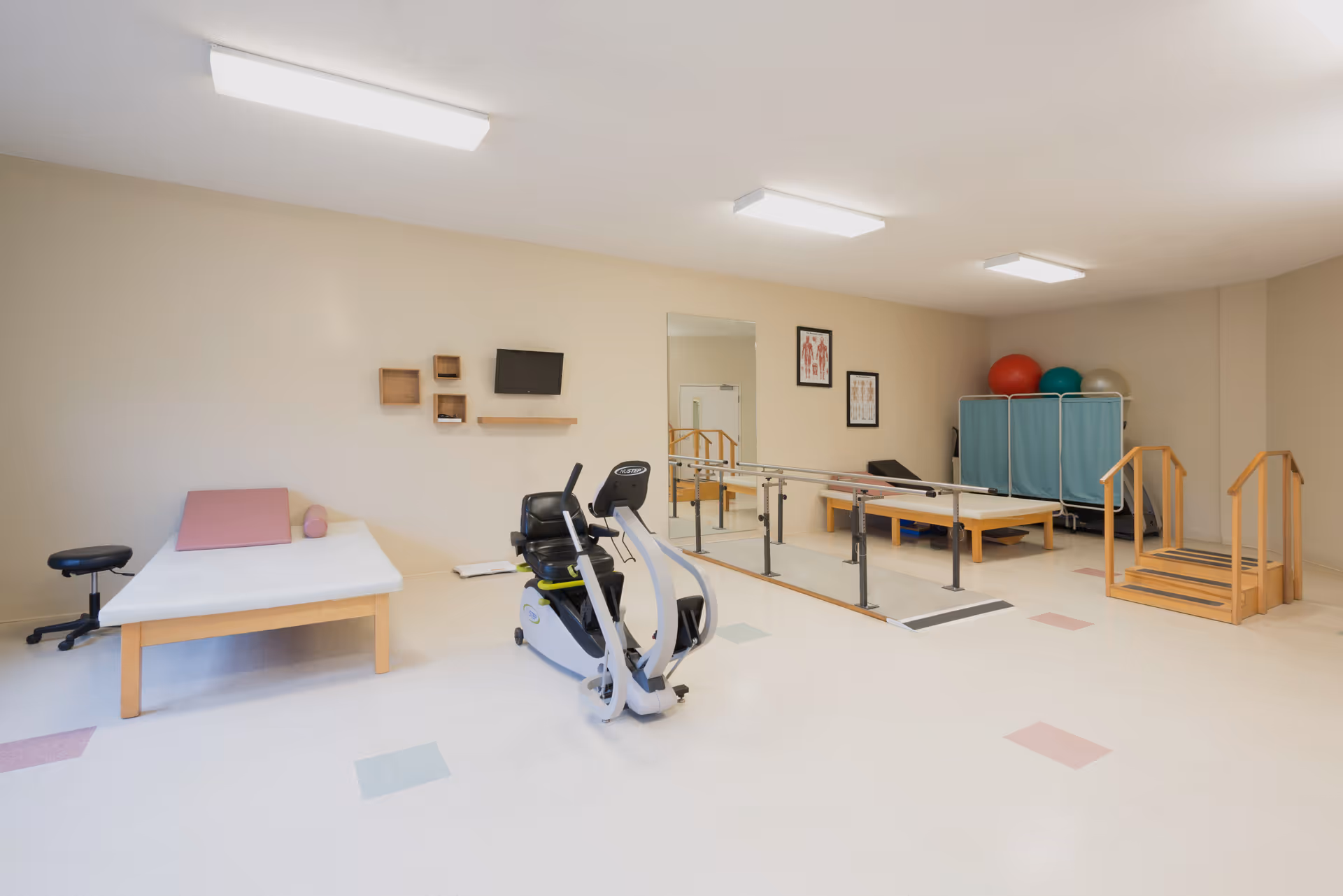 A physical therapy room with exercise equipment including a stationary recumbent bike, parallel bars for walking practice, a wooden step platform, a treatment table with a pink wedge cushion, anatomical charts on the wall, and a large mirror.