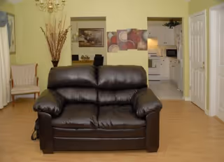 A cozy living room area featuring a dark brown leather loveseat centered on a wooden floor. Behind the loveseat, there is a wall with two open windows showing a dining area with a table and chairs, and a kitchen with white cabinets and appliances. The walls are painted light yellow, and there is a decorative tall vase with dried branches on the left side along with a beige upholstered chair.