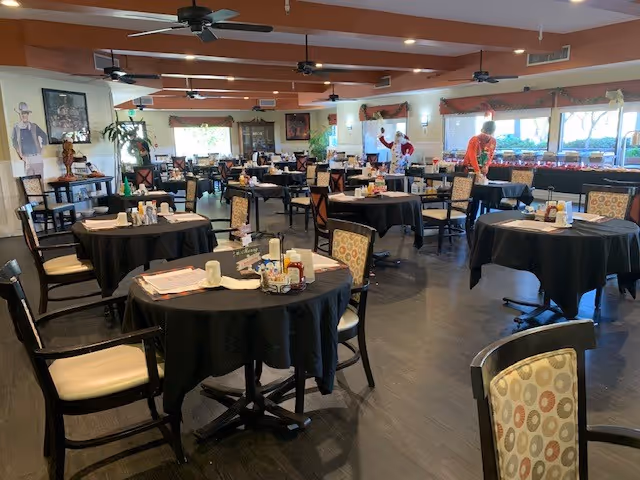 A spacious dining room with multiple round tables covered in black tablecloths, each set with condiments and napkins. Several chairs with patterned cushions surround the tables. In the background, two people are seen near a buffet area with large windows letting in natural light. Ceiling fans and recessed lighting are visible on the ceiling.