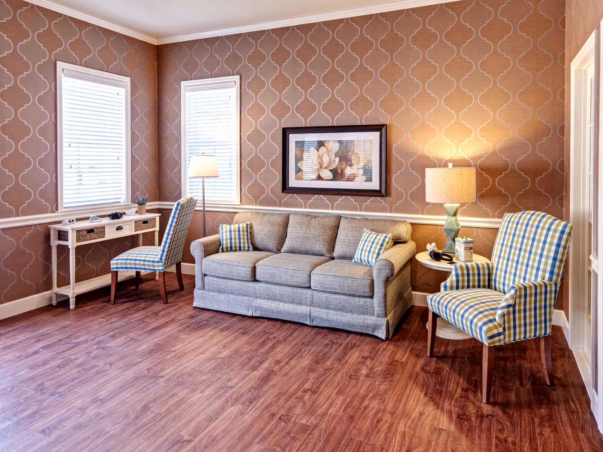 A cozy living room with patterned brown wallpaper, two windows with white blinds, a gray sofa with two plaid pillows, a plaid armchair, a small white side table with a lamp and decorative items, a white desk with a plaid chair, and wooden flooring.