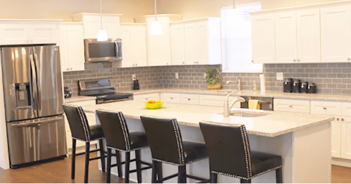 Modern kitchen with white cabinets, gray subway tile backsplash, stainless steel refrigerator, stove, and microwave. A large island with a granite countertop features a sink and four black bar stools with nailhead trim. Pendant lights hang above the island.