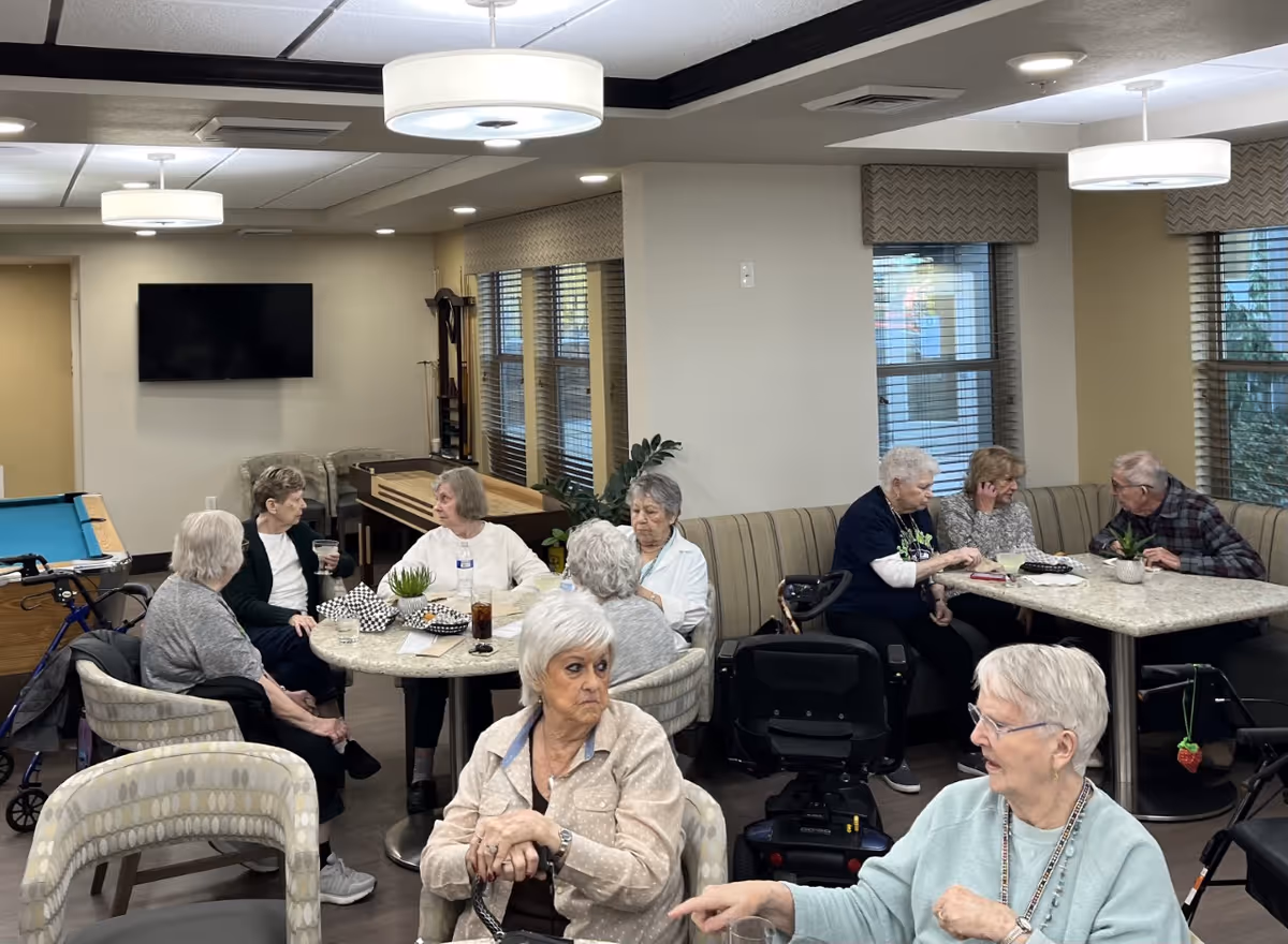 A group of elderly people sitting and socializing around tables in a well-lit common area with large windows, modern light fixtures, and a pool table in the background.