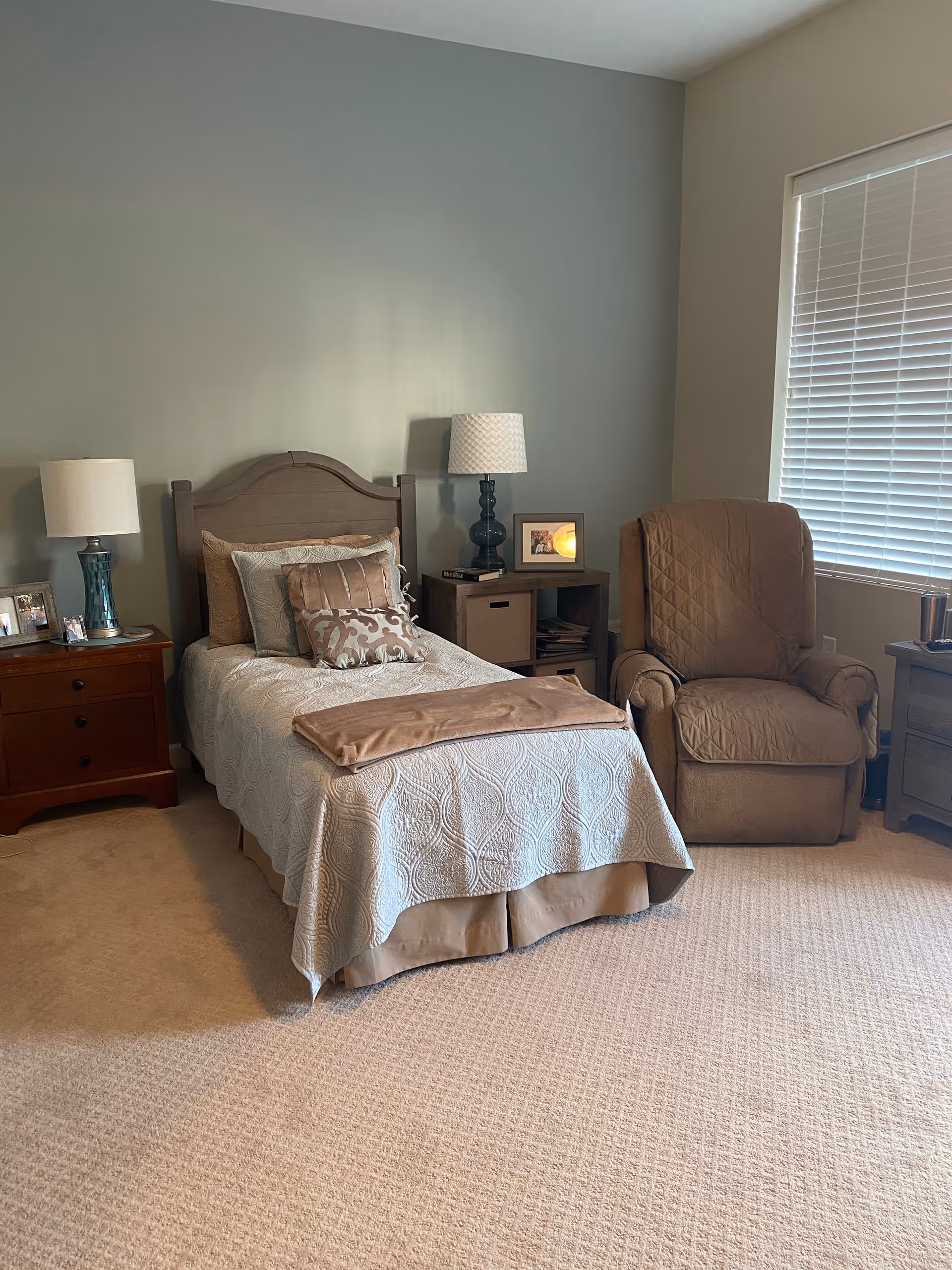 A cozy bedroom with a single bed covered in a beige quilt and multiple pillows. There is a wooden nightstand with a lamp and framed photos on the left side of the bed. On the right side, there is a small shelving unit with a lamp and framed picture on top, and a comfortable beige recliner chair next to a window with closed blinds. The room has beige carpeting and light-colored walls.