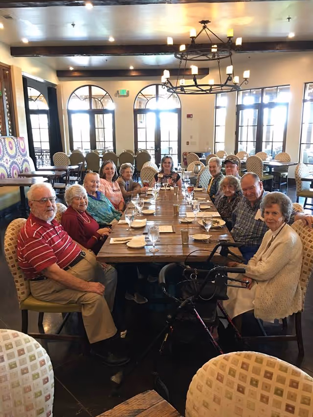 Elderly residents seated around a long wooden dining table in a bright dining room with arched windows.