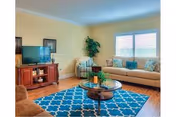 A cozy living room with a beige sofa, a patterned armchair, a wooden TV stand with a flat-screen television, a round glass coffee table on a blue patterned rug, and a large window with blinds letting in natural light.
