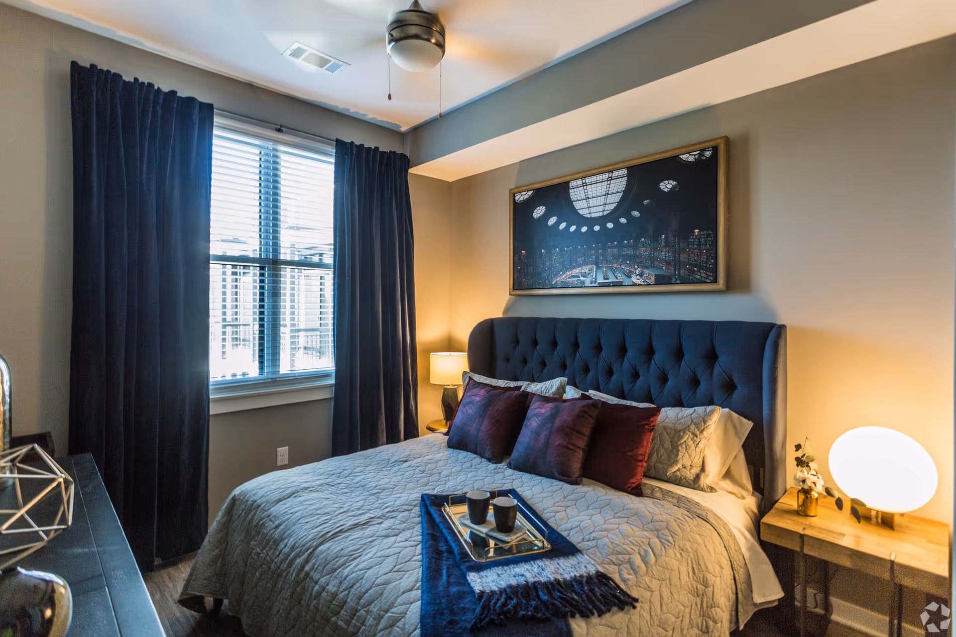A cozy bedroom with a large bed featuring a tufted dark blue headboard, beige quilted bedding, and several dark red and beige pillows. A blue throw blanket with a tray holding two black cups is placed on the bed. There is a window with closed blinds and dark curtains on the left wall, a ceiling fan above, a framed picture hanging above the bed, and two bedside tables with lamps and decorative items.
