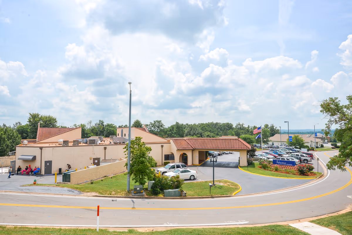 Exterior view of Signature HealthCARE of Cleveland facility showing a beige building with a red-tiled roof entrance, a parking lot with several cars, an American flag, and a blue facility sign. The scene includes a curved road in the foreground and trees in the background under a partly cloudy sky.