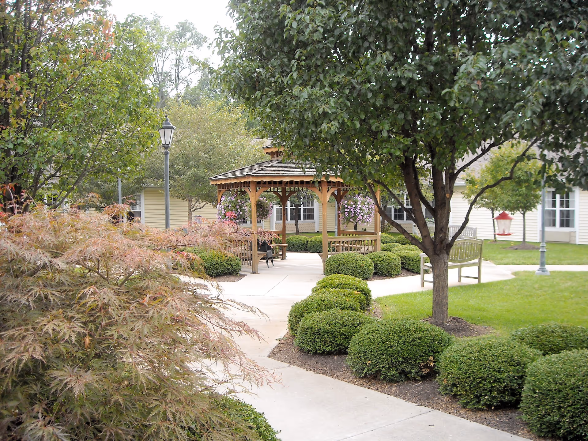 A landscaped outdoor garden area with a wooden gazebo surrounded by green bushes and trees. There are benches along the paved walkway, and hanging flower baskets decorate the gazebo. A building with windows is visible in the background.