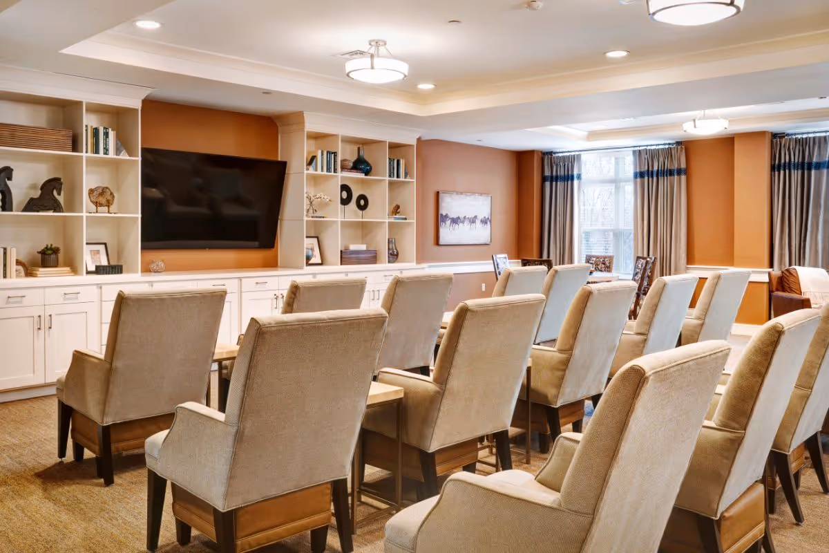 A cozy common area with multiple beige upholstered armchairs arranged in rows facing a wall-mounted flat screen TV. The room features built-in white shelves with decorative items and books, warm brown walls, and large windows with curtains allowing natural light to enter.