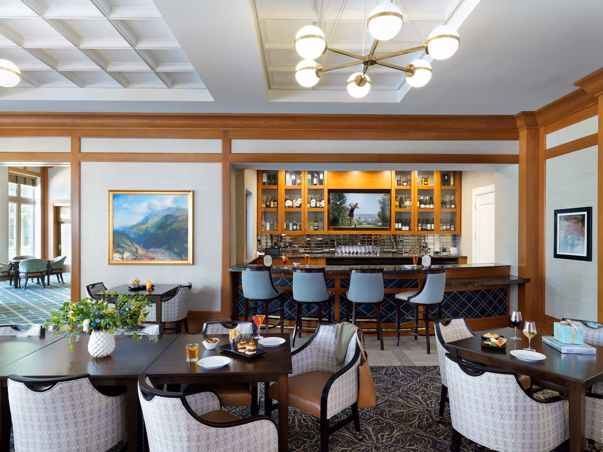 A stylish dining area with dark wooden tables and cushioned chairs arranged around them. A bar with four high chairs is visible in the background, with shelves holding bottles and glasses. A flat-screen TV above the bar shows a golfer in mid-swing. The room features wood paneling, a coffered ceiling with modern light fixtures, and framed artwork on the walls. Some tables have drinks, plates, and a flower arrangement.