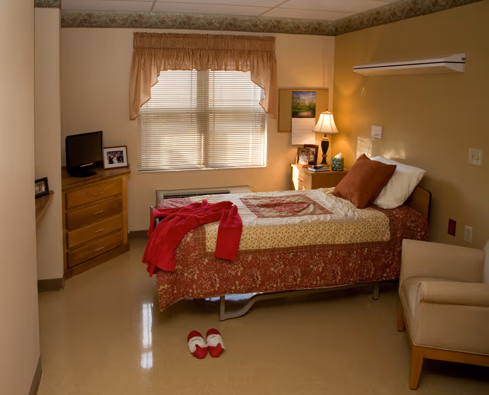 A cozy bedroom in a senior living facility with a single bed covered in a patterned quilt and a red blanket. There is a window with blinds and a valance above the bed. A wooden dresser with a small TV and framed photos sits to the left, and a nightstand with a lamp, calendar, and more framed photos is on the right. A pair of red slippers is placed on the floor near the foot of the bed, and a beige armchair is positioned in the corner.