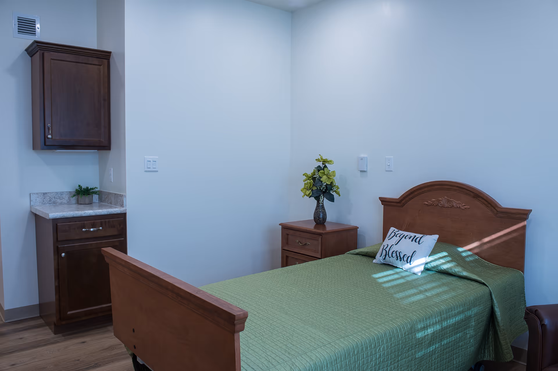 A simple bedroom with a wooden bed frame and green bedspread. There is a pillow on the bed with the words 'Beyond Blessed'. Next to the bed is a wooden nightstand with a vase of yellow flowers. On the left side of the room, there is a small countertop with a cabinet above and below it. The walls are plain white and the floor is wooden.